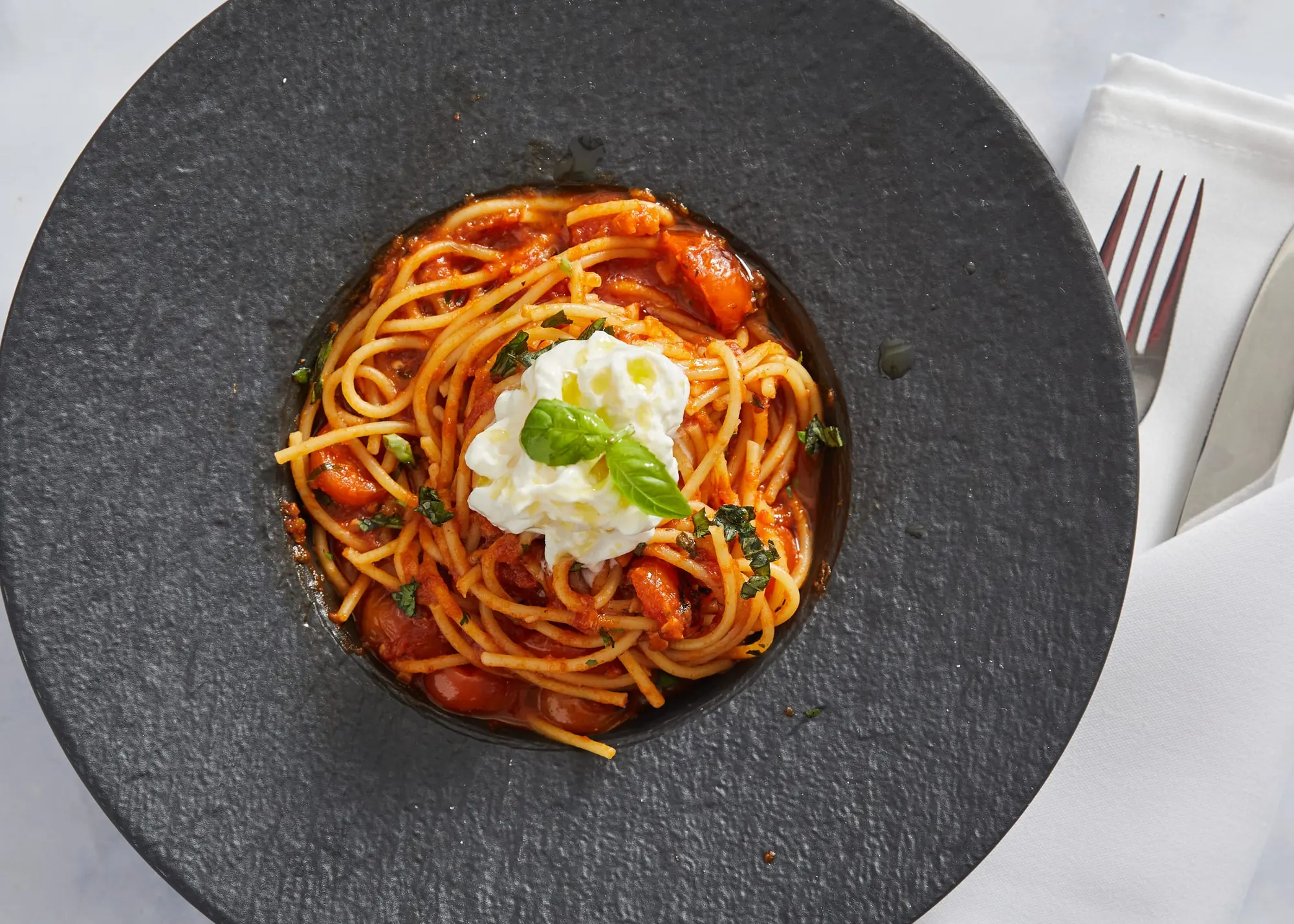 Spaghetti pomodori topped with burrata and basil in a dark bowl